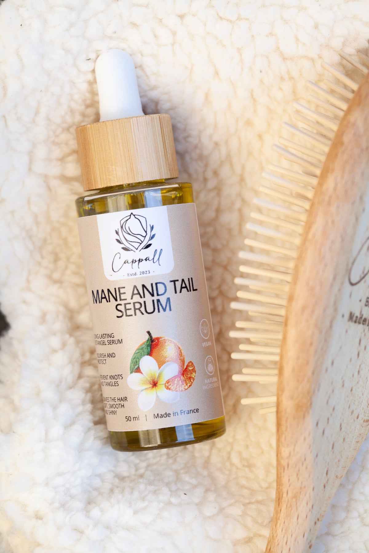 A bottle of Capell Mane and Tail Serum with a bamboo dropper cap, resting on a textured cream towel next to a wooden hair brush.