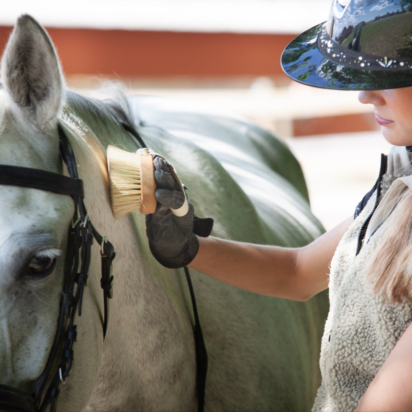 A woman in equestrian attire, including a black riding helmet and a textured vest, brushes a white horse's neck with a soft bristle. The scene is set in an equestrian stable with a warm, blurred background.