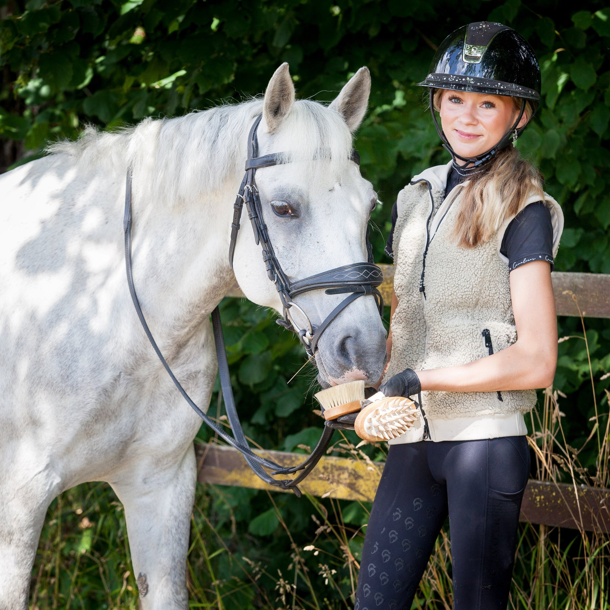 A woman in equestrian attire, including a black riding helmet, black gloves, and a beige vest, stands next to a white horse outdoors, holding a horse grooming kit for the horse.